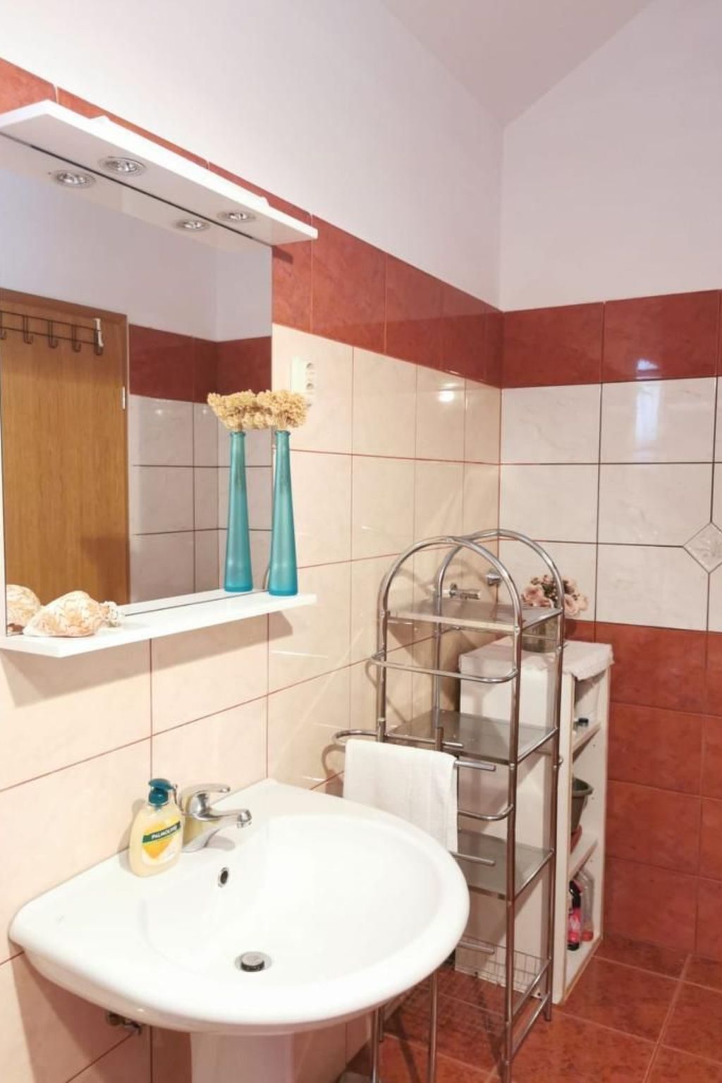 Un baño con lavamanos blanco, un espejo, toallero y estante con artículos de aseo.