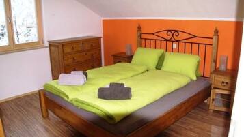 Un lit simple avec un cadre en bois, une couvre-lit verte et deux oreillers. Il y a une commode en bois sur la gauche et une table de chevet avec une lampe sur la droite.