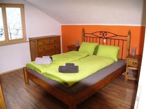 A single bed with a wooden frame, a green bedspread, and two pillows. There is a wooden dresser to the left and a nightstand with a lamp to the right.