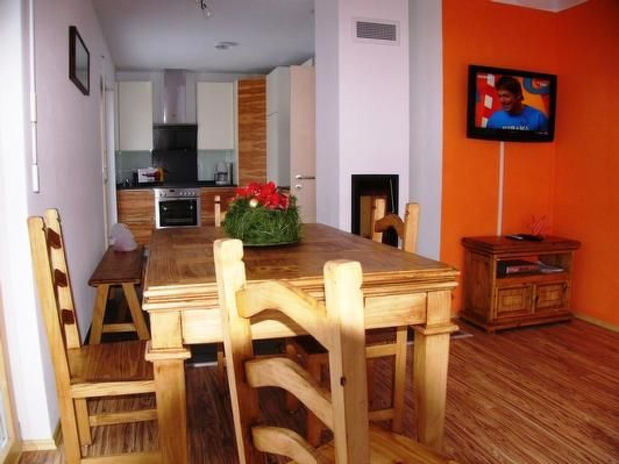 Un coin repas avec une table et des chaises en bois, une cuisine à l’arrière-plan et un téléviseur accroché au mur.