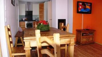 Un coin repas avec une table et des chaises en bois, une cuisine à l’arrière-plan et un téléviseur accroché au mur.
