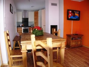 A dining area with a wooden table and chairs, a kitchen in the background, and a television on the wall.