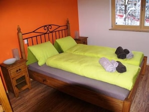 A bed with a wooden frame, a green bedspread, and pillows. There is a nightstand with a lamp and a small drawer.