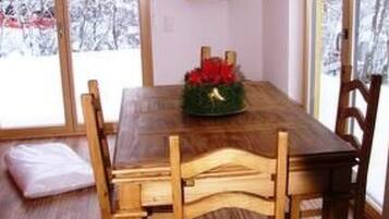 Une salle à manger avec une table et des chaises en bois, une petite étagère remplie de livres et une fenêtre donnant sur des arbres couverts de neige.