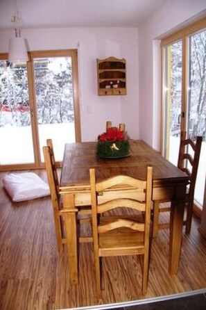 A dining room with a wooden table and chairs, a small shelf with books, and a window with a view of snow-covered trees.
