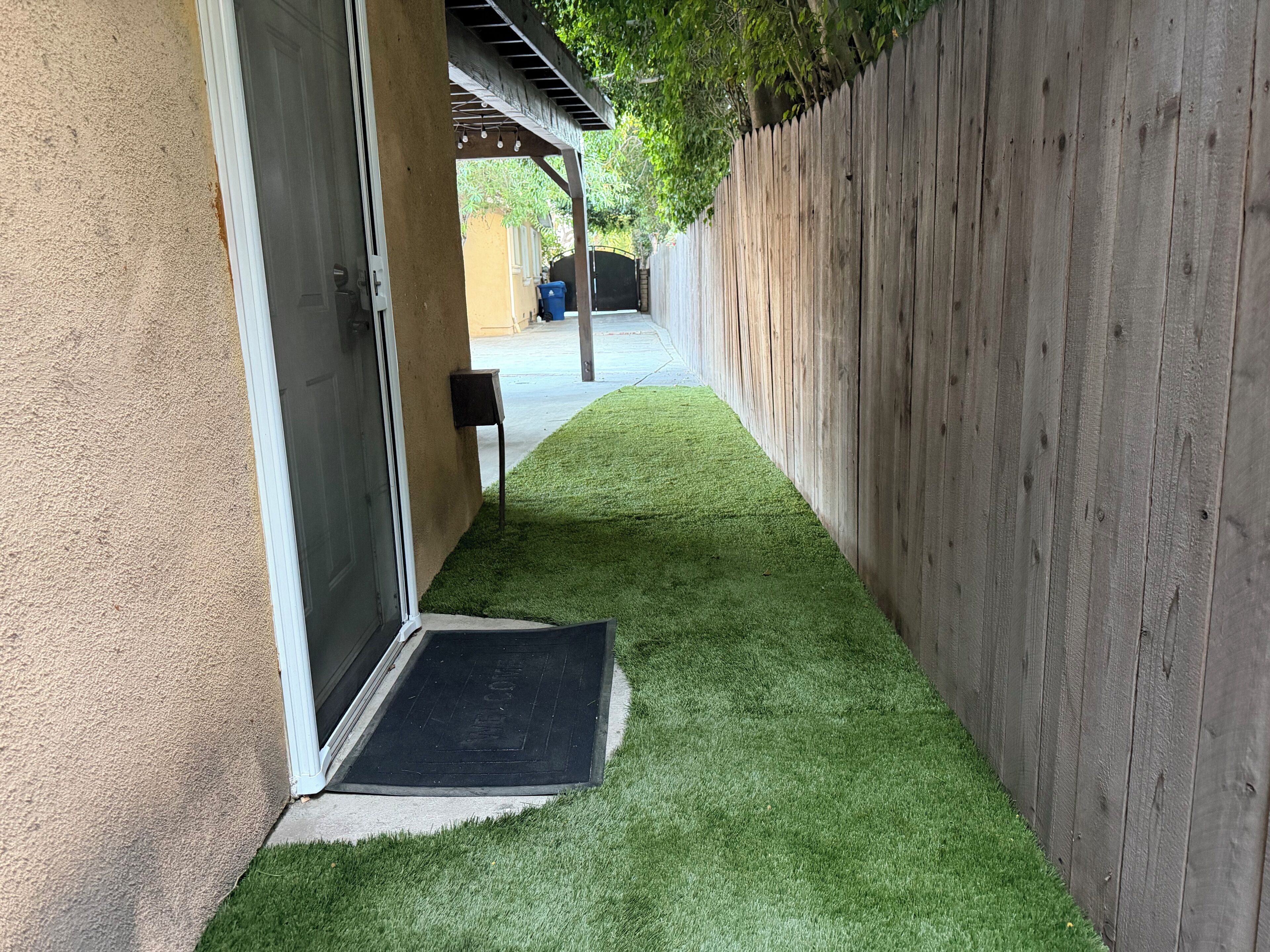 Un étroit sentier recouvert d’herbe artificielle mène à une porte, de part et d’autre de laquelle se trouvent une clôture en bois et un mur d’immeuble.