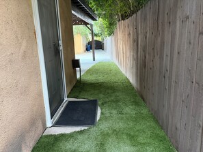 Un étroit sentier recouvert d’herbe artificielle mène à une porte, de part et d’autre de laquelle se trouvent une clôture en bois et un mur d’immeuble.
