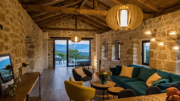 Interior rústico de piedra con techo de madera, un sofá, una mesa de comedor y vistas a través de un ventanal.