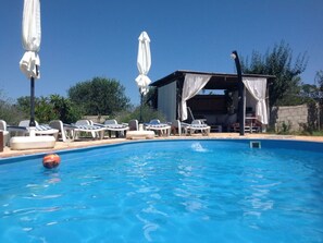 Une piscine avec des chaises longues et des parasols, un espace de détente couvert et un ciel d’un bleu limpide.