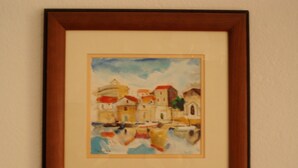 Pintura emoldurada retratando uma paisagem costeira com edifícios e barcos.