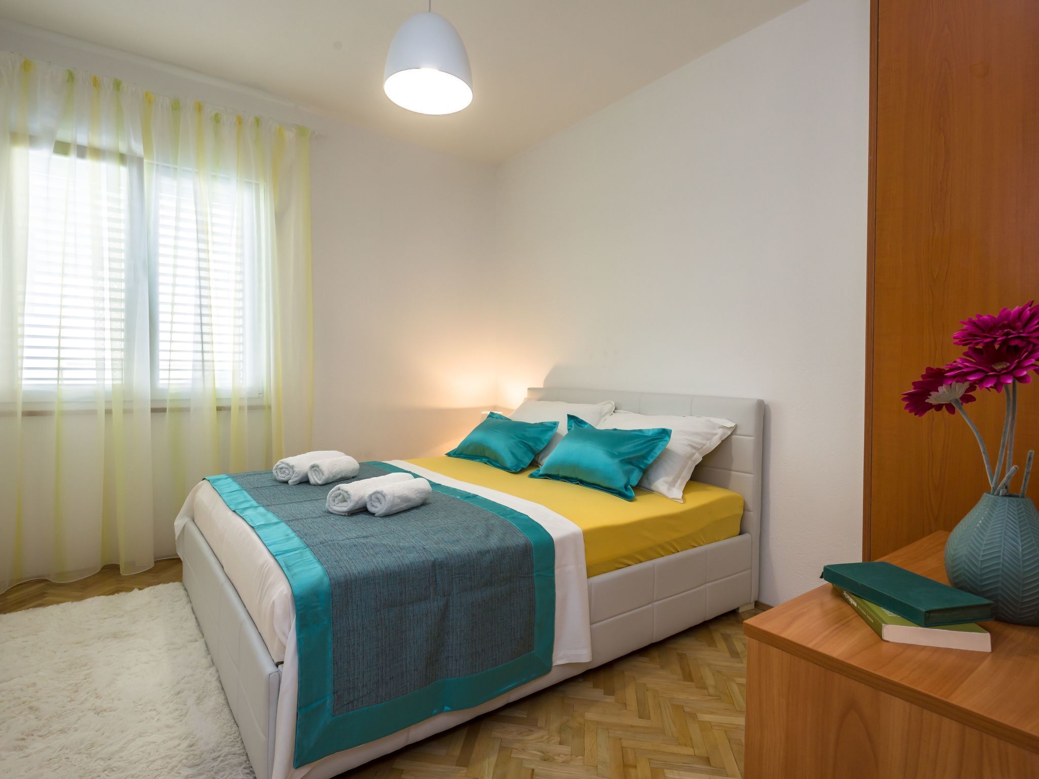 Ein ordentlich bezogenes Bett mit einer türkisfarbenen und weißen Bettdecke, ein Nachttisch aus Holz mit Büchern und einer Vase sowie ein Fenster mit transparenten Vorhängen.