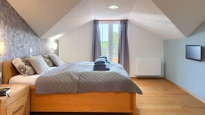 Un dormitorio con techo inclinado, una cama con colcha gris, una mesita de noche de madera y una ventana con cortinas.