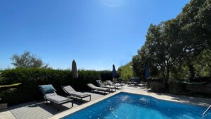 Piscina con sillones de descanso y sombrillas, rodeada de vegetación.
