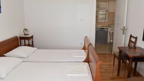 A room with two single beds, a wooden table, a chair, a small bedside table with a lamp, and a kitchen area visible through an open door.