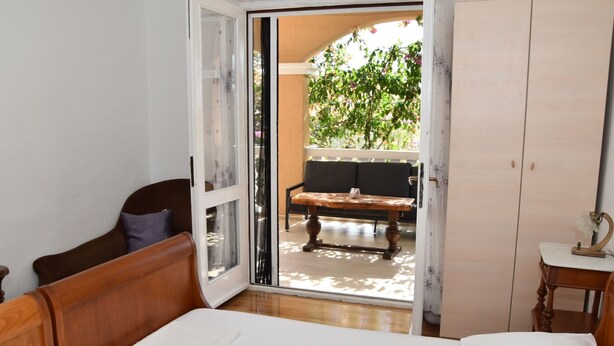 A bedroom with a bed, a wooden nightstand, a wardrobe, and a sliding glass door leading to a balcony with a bench and greenery.