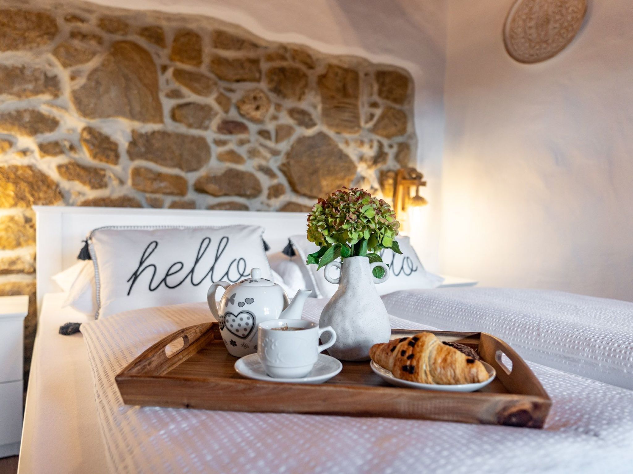 A neatly made bed with a wooden tray holding a teapot, a cup, and croissants. A vase with flowers is on the bed, and pillows with "hello" written on them are visible.