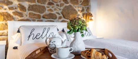 A neatly made bed with a wooden tray holding a teapot, a cup, and croissants. A vase with flowers is on the bed, and pillows with "hello" written on them are visible.
