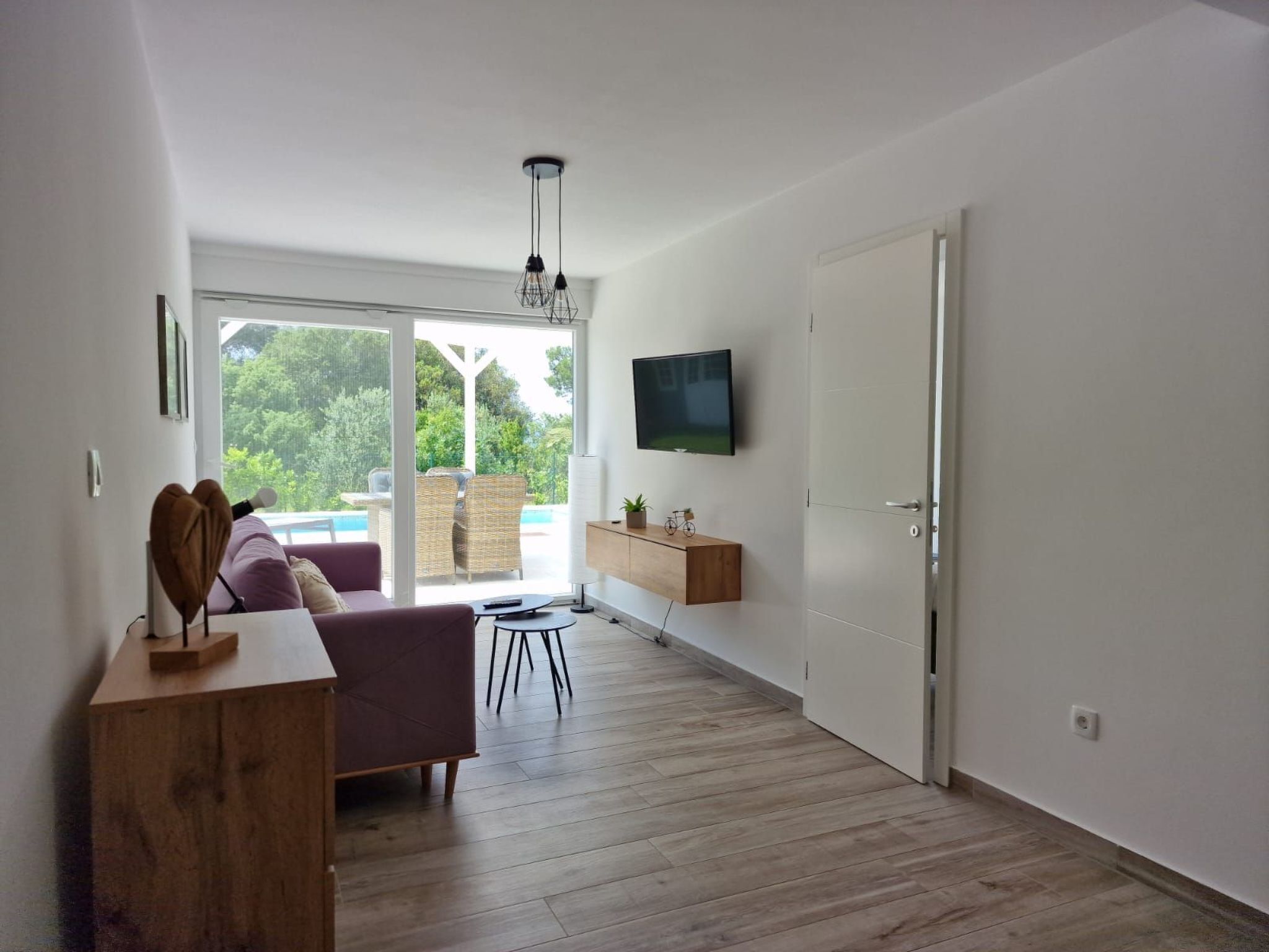 A modern living room with a large sliding glass door leading to a balcony, a flat-screen TV mounted on the wall, and a wooden coffee table.