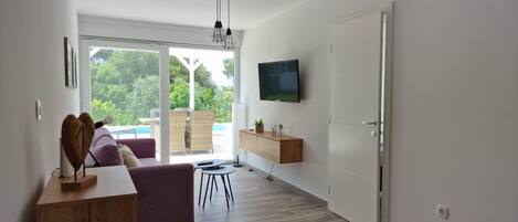 A modern living room with a large sliding glass door leading to a balcony, a flat-screen TV mounted on the wall, and a wooden coffee table.