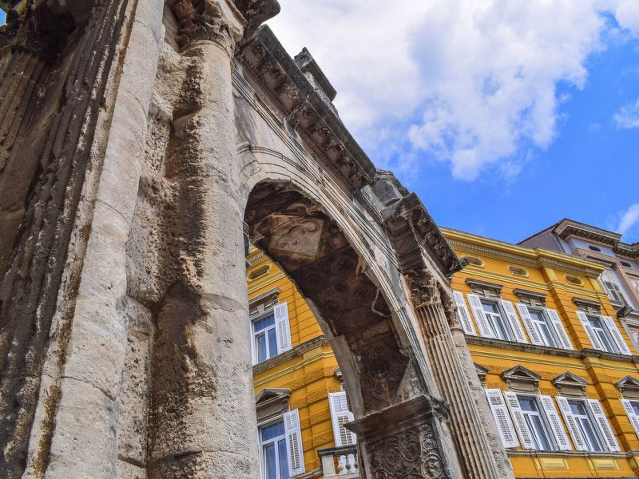 Um arco de pedra antiga com colunas desgastadas, conduzindo a um edifício amarelo com persianas brancas.