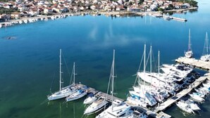 Uma marina com vários veleiros atracados, rodeada por uma vila costeira com casas e uma paisagem montanhosa ao fundo.
