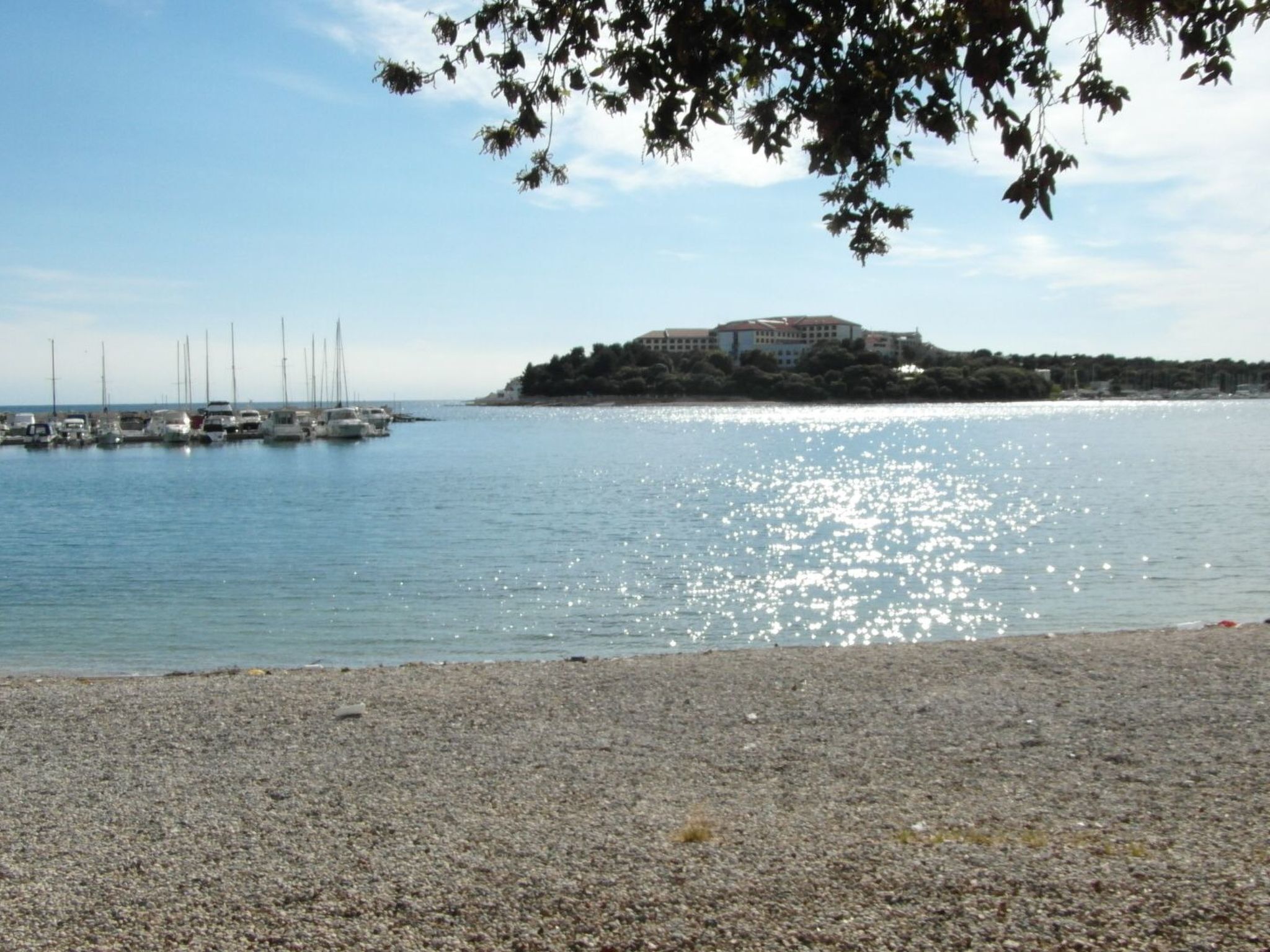 Escena costera con una playa pedregosa, un puerto deportivo con veleros y un edificio grande en una isla.