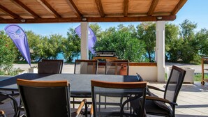 Eine Terrasse im Freien mit Holzbalken, Metallstühlen und einem Tisch. Im Hintergrund sind Flaggen und ein Grill zu sehen.