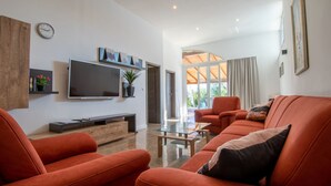 Ein modernes Wohnzimmer mit Fernseher, orangefarbenen Sofas, einem Glastisch und einem großen Fenster mit Vorhängen.