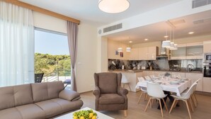Ein modernes Wohnzimmer mit einer Couch, einem Sessel und einem Esstisch für vier Personen. Ein großes Fenster bietet Blick auf Grünflächen.