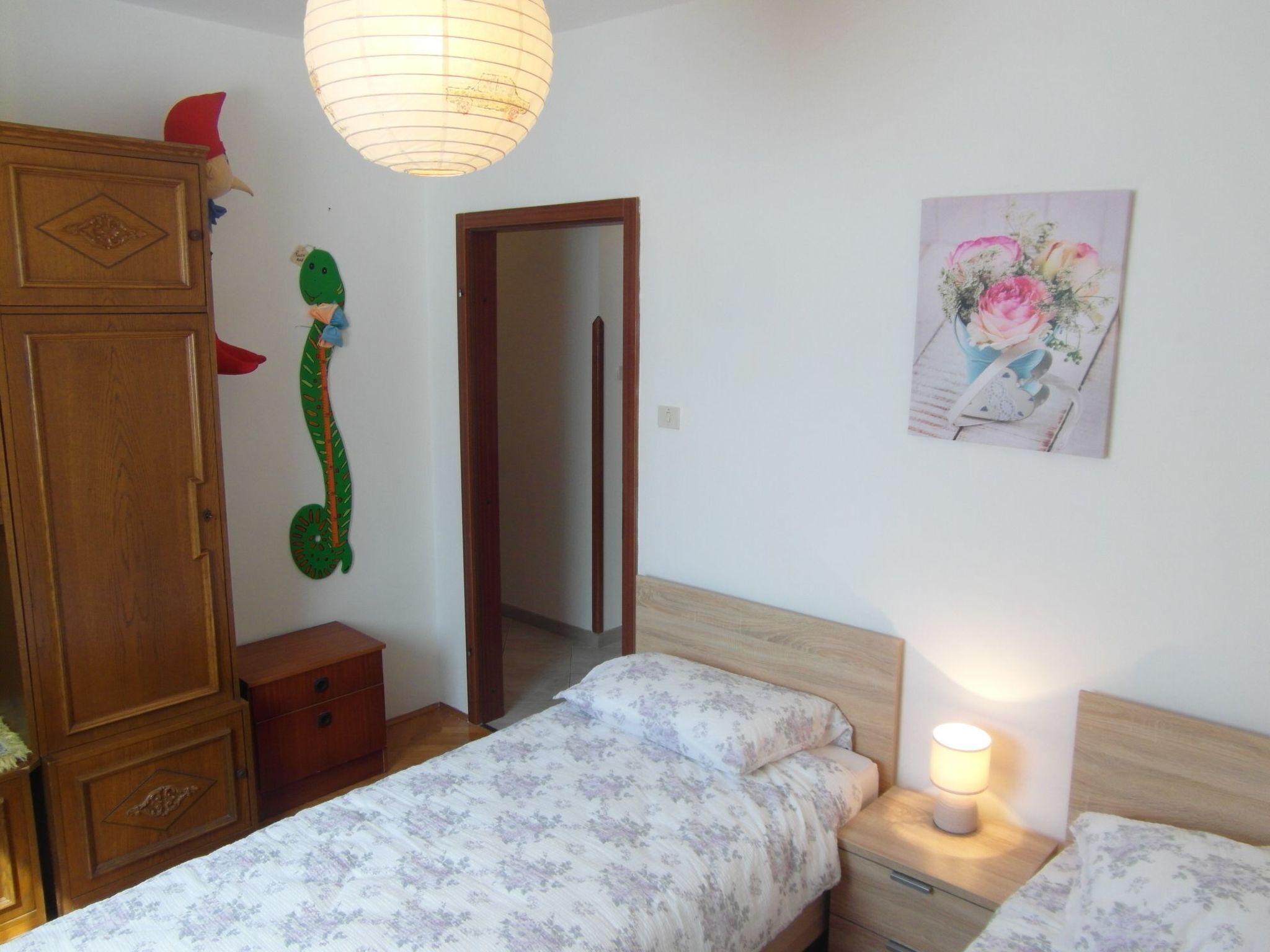A bedroom with two beds, a wooden wardrobe, a small bedside table with a lamp, and a door leading to another room.