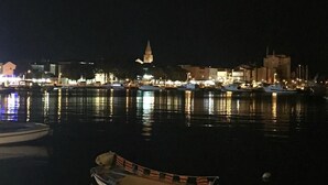 Vista noturna de um porto com barcos atracados, uma torre de igreja imponente e edifícios iluminados ao fundo.