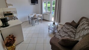 Una sala de estar moderna con un sofá, una mesa de centro y un comedor con mesa y sillas. Se observa una cocina con microondas y cafetera. Un televisor está montado en la pared, y se ve una ventana con cortinas.