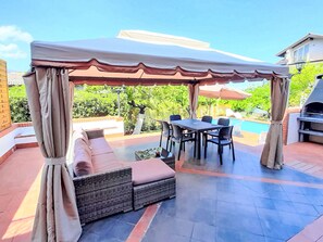 Eine Terrasse im Freien mit überdachter Sitzgruppe, einem Esstisch und einem Grill.