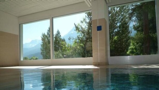 Ein Schwimmbecken mit klarem Wasser, großen Fenstern und Blick auf Bäume und Berge.