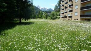 Ein Gebäude mit mehreren Balkonen, umgeben von einer grünen Wiese mit weißen Blumen und einem bewaldeten Gebiet im Hintergrund mit Bergen.