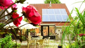 Una casa con techo solar, un patio con muebles de mimbre y un jardín con plantas y flores.