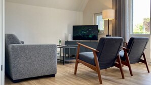 Un salon moderne avec un canapé gris, deux fauteuils, un téléviseur à écran plat, une petite table et une lampe.