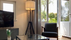 Un salon moderne doté d’un téléviseur à écran plat, d’une table basse en verre, d’un lampadaire, d’un fauteuil sombre et de grandes fenêtres donnant sur un paysage verdoyant.