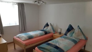 A small, simple room with two single beds, a bedside table with a lamp, and a window with curtains.