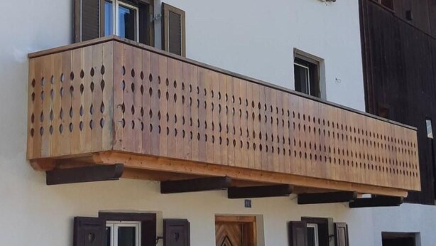 A building with a wooden balcony with circular cutouts, a door with a diamond pattern, and shutters on windows.