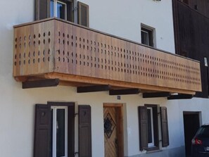 A building with a wooden balcony with circular cutouts, a door with a diamond pattern, and shutters on windows. - Wohnen im alten Bauernhaus,Wohnung EG by Interhome (Müstair)