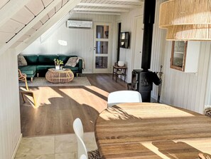 Ein gemütliches Wohnzimmer mit einem grünen Sofa, einem runden Holztisch und einem weißen Sessel. In der Ecke ist ein schwarzer Kaminofen zu sehen.