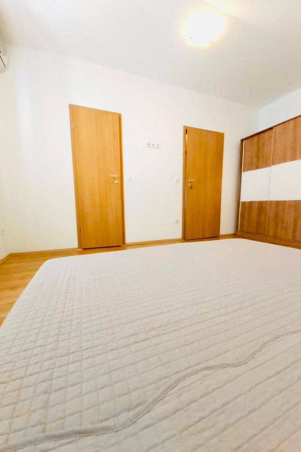 Une pièce avec un grand lit, deux portes en bois et un luminaire blanc au plafond.