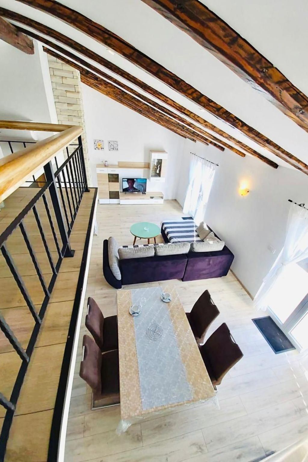 A spacious living area with a high ceiling, exposed wooden beams, a dining table with chairs, a sofa set, and a television.