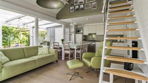 En kompakt opholdsstue med en grøn sofa, et spisebord og et tekøkken. En hvid trappe med kradsetræ er synlig.