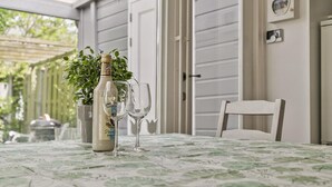 Et bord med dug, en flaske og to glas på en terrasse med en hvid dør og en plante i baggrunden.