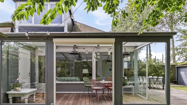 Et moderne hus med en glasindrammet patio med spisebord og stole, omgivet af en træterrasse.