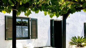 Ein Tisch unter einer Pergola, im Hintergrund eine weiße Wand und grüne Fensterläden.