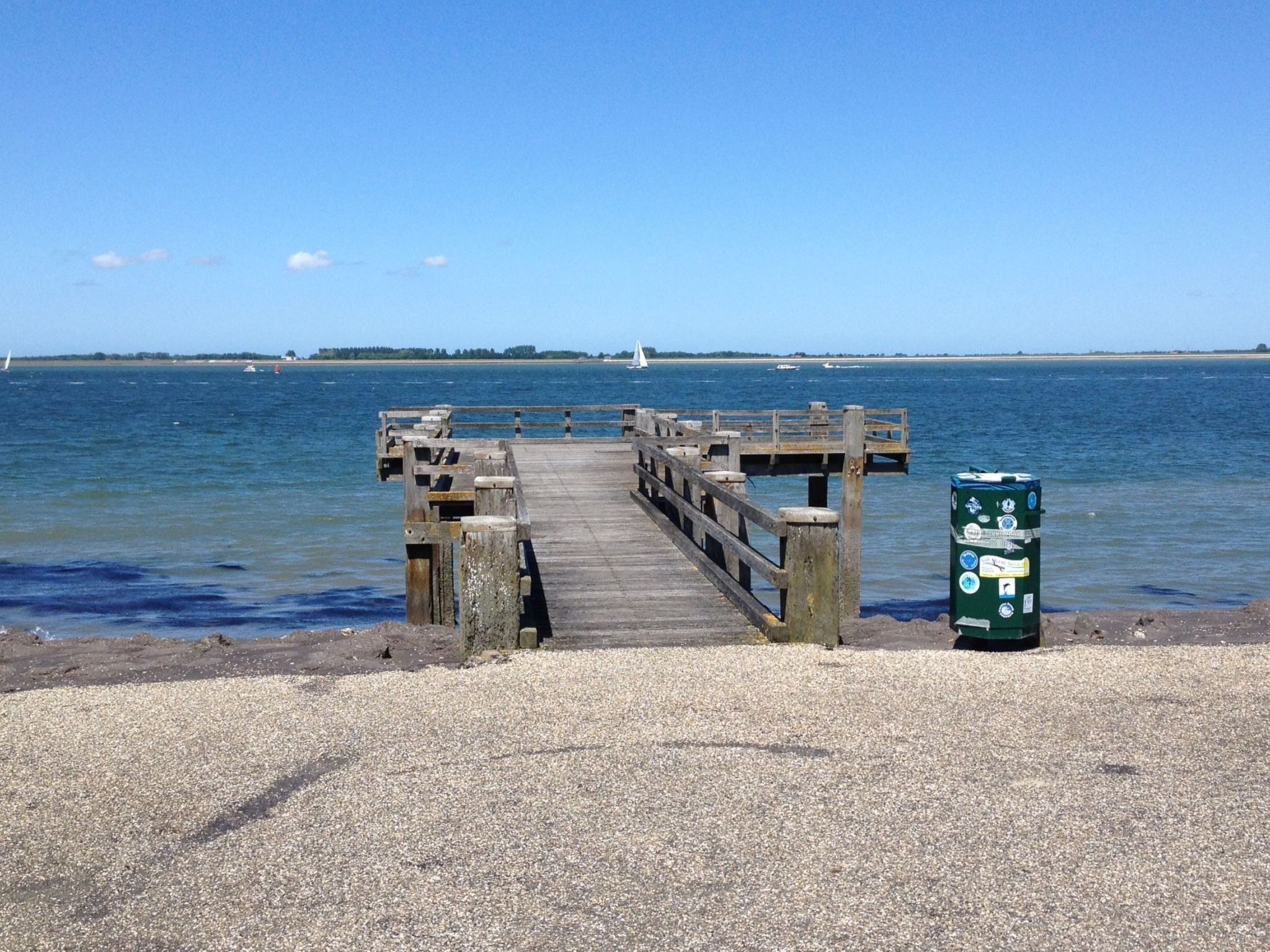 A wooden pier extending into calm waters with a green trash bin on the shore.