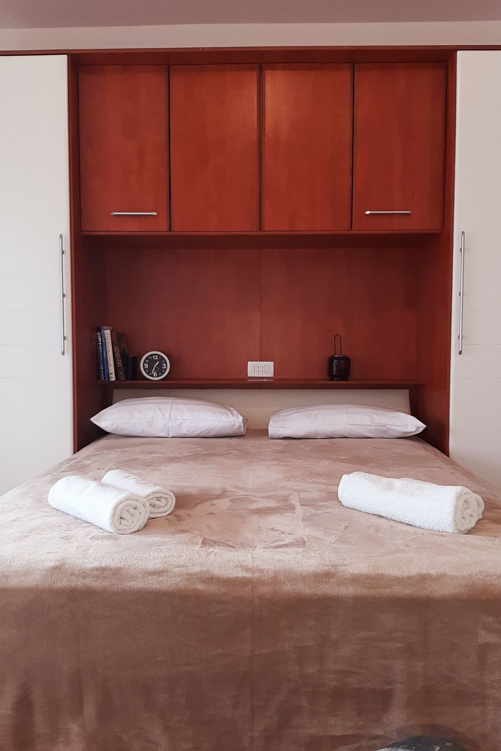 A bedroom with a bed, pillows, a clock, a power outlet, and a small speaker.